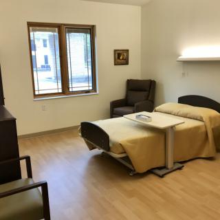 A neatly arranged hospital room with a single bed, chairs, and a window allowing natural light.