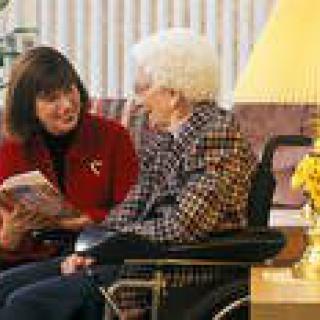 A caregiver reads to an elderly woman sitting in a wheelchair indoors.