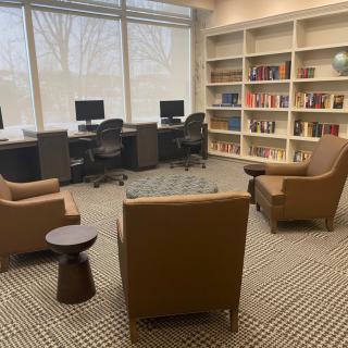 A cozy library reading area featuring comfortable chairs, bookshelves, and computer stations.
