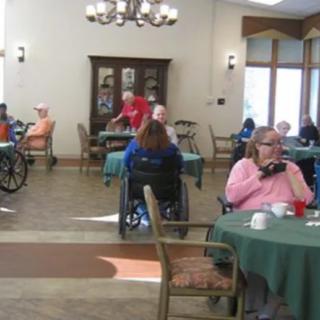 Residents in a nursing home dining area, some in wheelchairs, engaging in various activities.