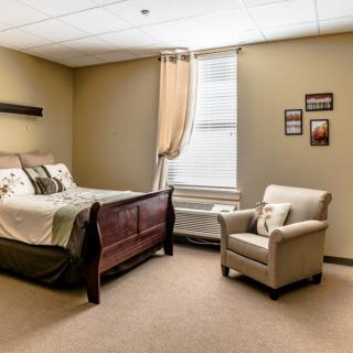 A cozy bedroom with a bed, an armchair, and a window, featuring soft lighting and simple decor.