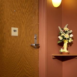 A cozy interior scene featuring a wooden door, a small shelf with a flower arrangement in a vase, and a wall-mounted light.