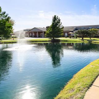 A picturesque view of a pond with a fountain, surrounded by trees and a building in the background.