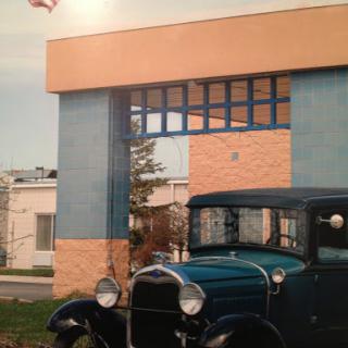 A vintage car parked in front of a historic building, with an American flag waving in the background.