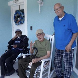 A group of three elderly men relaxing on a porch, with one in a wheelchair and another in a rocking chair.