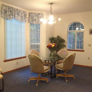 A cozy dining room with a glass table and four cushioned chairs, illuminated by a chandelier. The room features large windows with vertical blinds and patterned curtains.