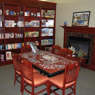 A cozy room featuring a table with a puzzle, surrounded by chairs, a bookshelf, and a fireplace.
