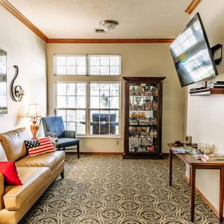 A cozy living room with a comfortable sofa and armchair, decorated with a patterned carpet and wall art. A large window lets in light, and a TV is mounted on the wall above a wooden cabinet displaying decorative items.