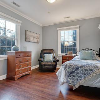 A cozy bedroom with hardwood floors, featuring a bed, dresser, and chair by large windows.