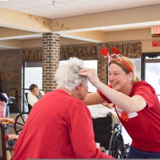 A caregiver joyfully interacts with an elderly resident in a community setting, both wearing red to celebrate a special occasion.