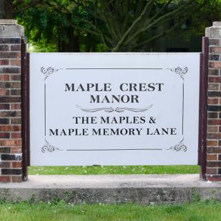 A sign for Maple Crest Manor, with mentions of The Maples and Maple Memory Lane, surrounded by greenery.