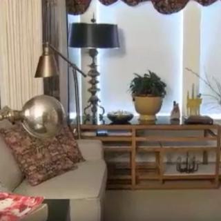 A cozy living room with a sofa, decorative pillows, a table lamp, and plants on the coffee table in front of window blinds.