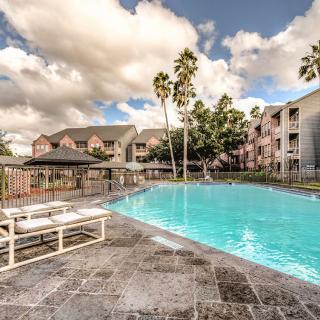 A serene apartment complex featuring a sparkling swimming pool surrounded by palm trees and buildings under a partly cloudy sky.