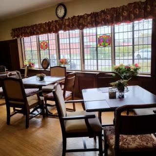 A cozy dining area with neatly arranged tables and chairs, decorated with flowers and illuminated by natural light through large windows.