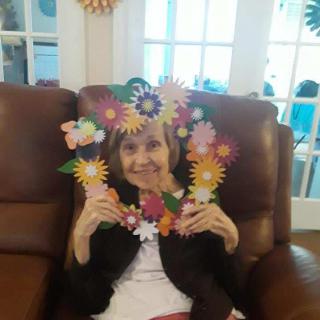 An elderly woman smiling while holding a colorful flower frame in a living room.