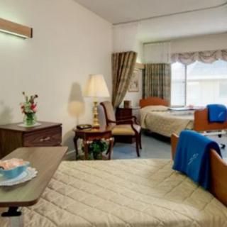 A hospital room with two beds, featuring a nightstand, lamp, and a window with curtains.