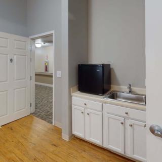A small kitchenette area with white cabinetry, a television, and hardwood flooring, leading to an adjacent room through an open doorway.
