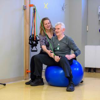 A therapist assists an elderly man in exercises using a blue exercise ball in a rehabilitation setting.