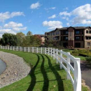 Scenic view of a white fence curving around a pond, with apartment buildings in the background and a blue sky above.