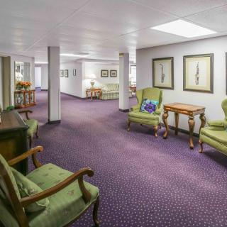 A well-lit hallway with green upholstered chairs and wooden tables, featuring framed pictures on the walls.