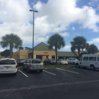 A parking lot with several cars and a building in the background, surrounded by palm trees under a cloudy sky.