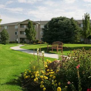 A landscaped garden area with blooming flowers, trees, a bench, and an apartment building in the background.