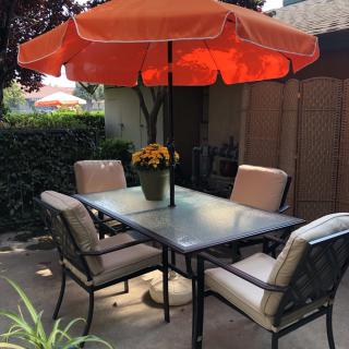 A cozy patio setup with a glass table, cushioned chairs, and an orange umbrella providing shade.