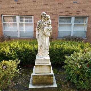 A statue stands in front of a brick building surrounded by bushes.