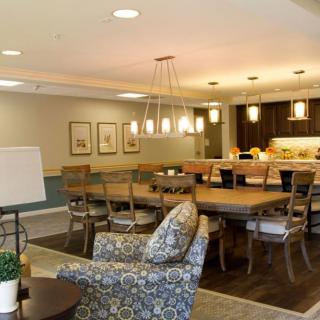 A modern dining area with a large wooden table and a cozy kitchen in the background, featuring stylish lighting and comfortable seating.