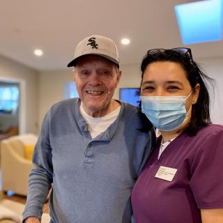 An elderly man smiling next to a caregiver wearing a mask indoors.