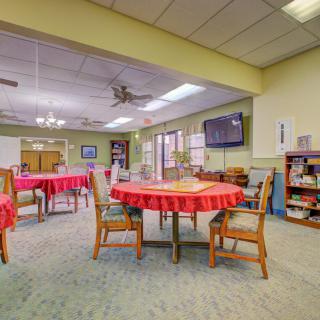 A spacious dining room with tables covered in red tablecloths, surrounded by wooden chairs, featuring a television and bookshelves on the walls.