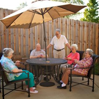 A group of seniors enjoys a relaxed conversation around a patio table under an umbrella in a garden setting.