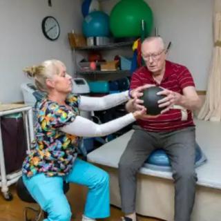 A healthcare professional assists an elderly man in a physical therapy session using a medicine ball.