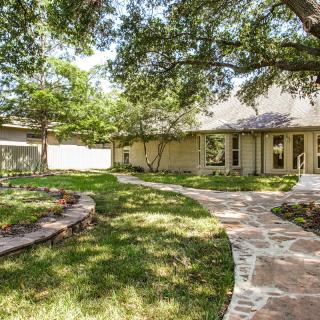 A charming house with a stone pathway winding through a well-maintained garden and lawn, shaded by large trees.