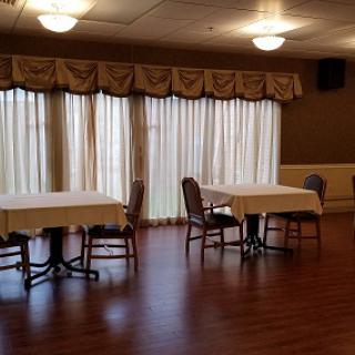 An interior view of a room with tables and chairs, featuring large windows draped with curtains.