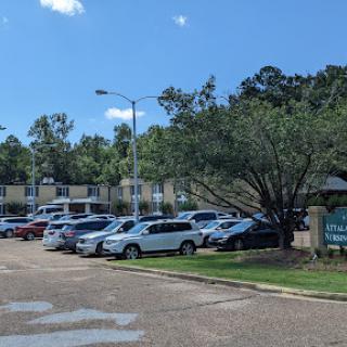 A busy parking lot in front of an apartment complex, surrounded by trees under a partly cloudy sky.