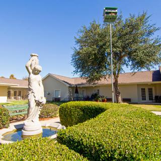 A serene garden scene featuring a classical statue, neatly trimmed bushes, and a single-story house in the background under a clear blue sky.