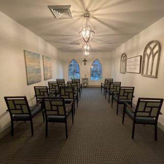 A small, symmetrical chapel with rows of chairs and a cross at the front, illuminated by soft lighting.