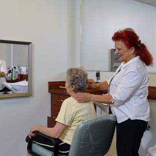 A hairdresser styling a senior woman's hair in a salon, with a mirror reflecting their image.