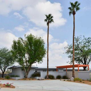A mid-century modern house with tall palm trees set against a partly cloudy sky in a desert landscape.