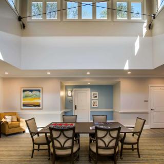 A modern living room with a central table surrounded by chairs under large windows.