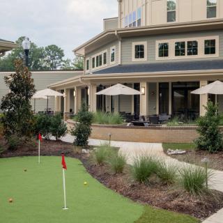 A view of a building with an outdoor patio featuring umbrellas and a golf putting green surrounded by landscaping.