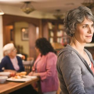 A woman stands in the foreground of a cozy kitchen while two elderly women engage in conversation over a meal.