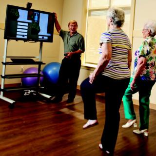 A group of senior citizens participating in a fitness class, following exercises displayed on a television screen indoors.