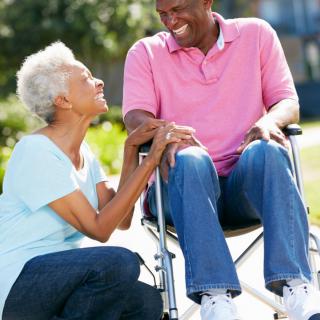 An elderly couple enjoys a sunny day outside, with one sitting in a wheelchair and both sharing a smile.