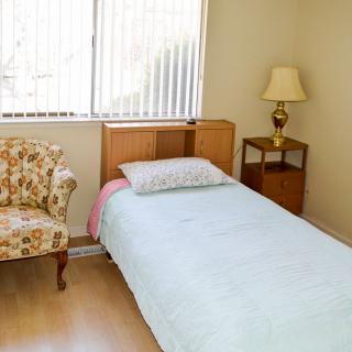 A simple bedroom with a single bed, floral chair, and a lamp on a nightstand by the window.