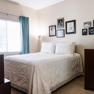 A cozy bedroom with framed artwork above the bed and light streaming through a window with blue curtains.