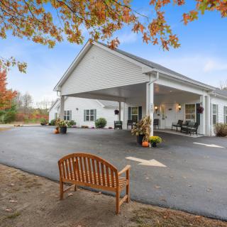 A building with a covered entrance, surrounded by autumn trees and a wooden bench in the foreground.