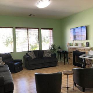 A cozy living room with green walls, featuring comfortable sofas, a television, and large windows letting in natural light.