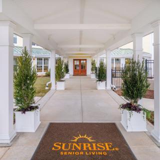 A covered walkway with potted plants leads to a building entrance, featuring a mat with the 'Sunrise Senior Living' logo.